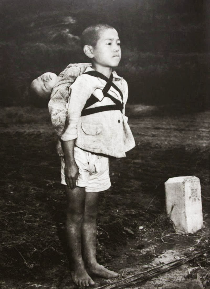 A Japanese boy standing at attention after having brought his dead younger brother to a cremation pyre, 1945